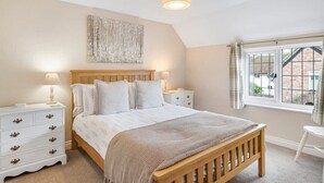A bedroom with a wooden bed, white nightstands, a bedside lamp, a window with curtains, and a wall-mounted artwork. - Idyllic Cottage in Heart of Devon Countryside (Cullompton)