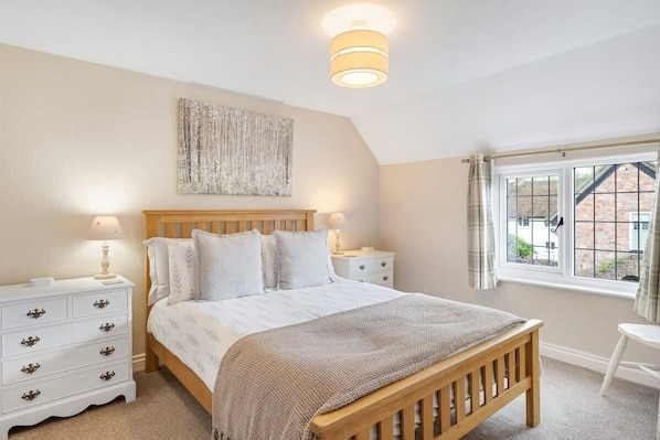 A bedroom with a wooden bed, white nightstands, a bedside lamp, a window with curtains, and a wall-mounted artwork. - Idyllic Cottage in Heart of Devon Countryside (Cullompton)