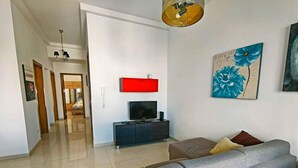 A modern living room with a grey sofa, a flat-screen TV on a black stand, and a red accent wall. There is a painting of blue flowers on the wall and a hallway leading to another room. - Remarkable 2-bed Apartment in St Julians (St. Julian's)