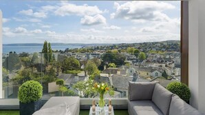 Un salon moderne doté d’une cloison en verre, d’un canapé gris, d’une petite table sur laquelle est posé un vase de fleurs, et offrant une vue sur un paysage urbain et sur l’eau.