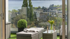 Un balcon donnant sur un lac et des bâtiments, un canapé en osier, une petite table avec des verres et des bouteilles, et une plante en pot.