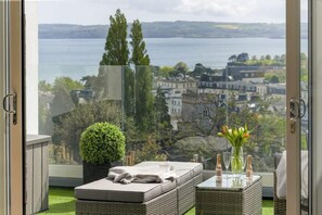 A balcony with a view of a lake and buildings, a wicker sofa, a small table with glasses and bottles, and a potted plant. - Apartment with Stunning Sea Views, Private Parking (Torquay)