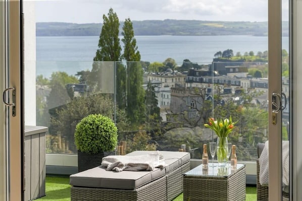 A balcony with a view of a lake and buildings, a wicker sofa, a small table with glasses and bottles, and a potted plant. - Apartment with Stunning Sea Views, Private Parking (Torquay)