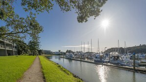 A marina with boats docked along a calm waterway, a grassy path beside the water, and buildings in the background.