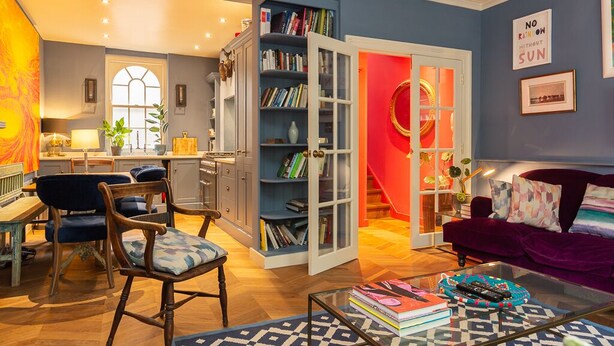 Ein gemütliches Wohnzimmer mit einer roten Tür, die zur Küche führt, einem violetten Sofa und einem Bücherregal voller Bücher.