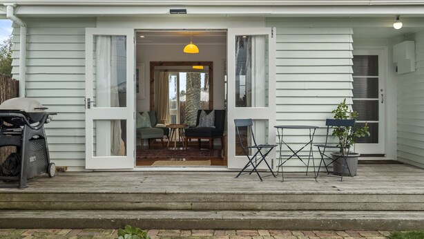 Een huis met een open voordeur, waarachter een woonkamer zichtbaar is met een bank, een kleine tafel en een stoel. Buiten de deur staat een barbecue, en op de veranda een bloempot.