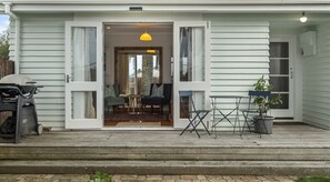 Ein Haus mit offener Haustür, aus der ein Wohnzimmer mit Sofa, einem kleinen Tisch und einem Stuhl zu sehen ist. Vor der Tür steht ein Grill und auf der Veranda ein Blumentopf.