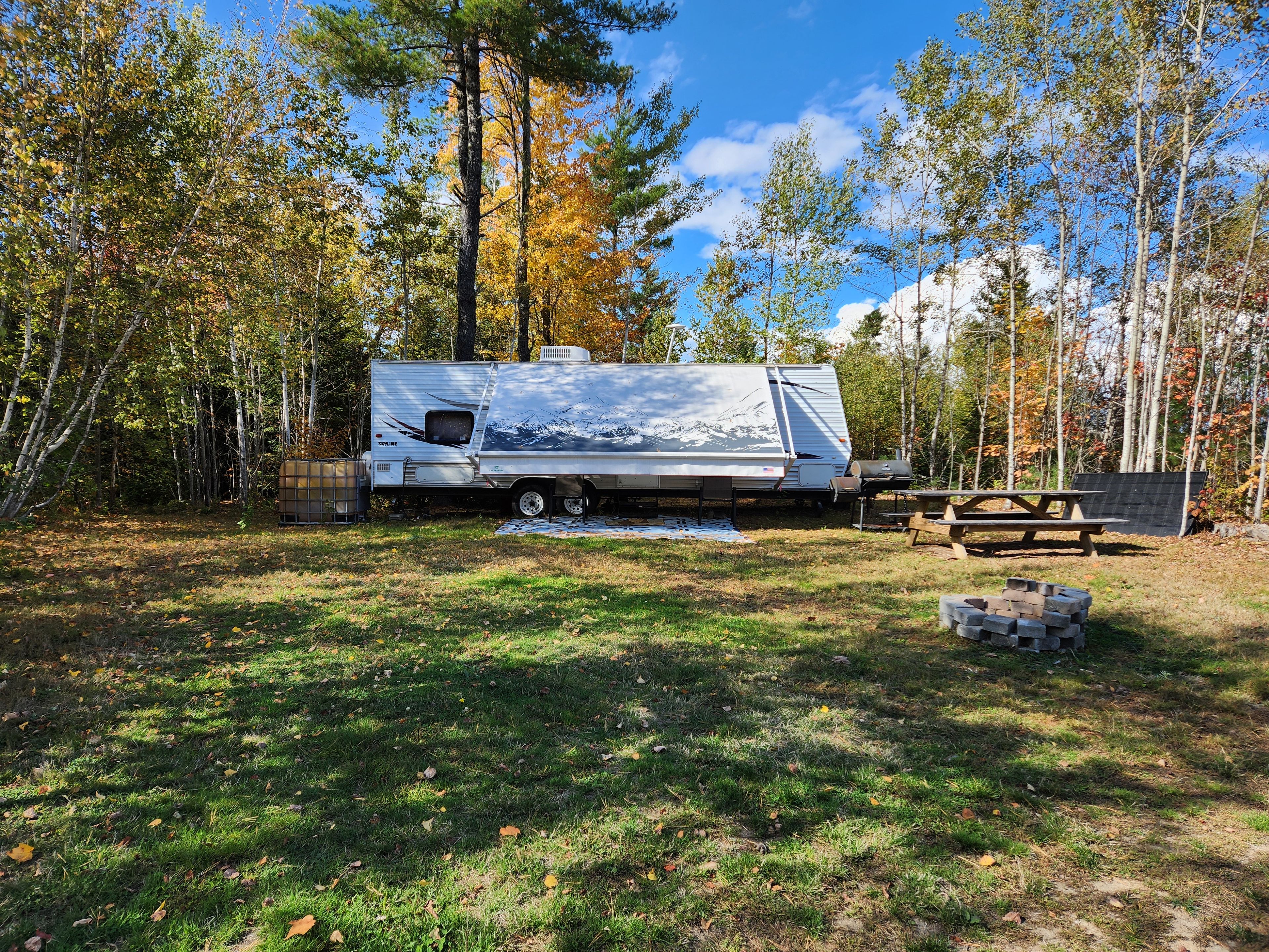 A recreational vehicle (RV) parked in a wooded area with a picnic table and a grill nearby.