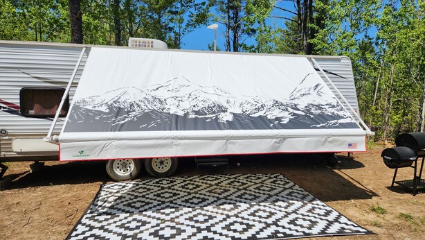 A travel trailer with a mountain design on the side, a patterned rug on the ground, and trees in the background.