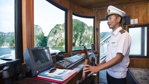 Een scheepvaartcontrolekamer met een kapitein, navigatieapparatuur en uitzicht op zee en bergen.