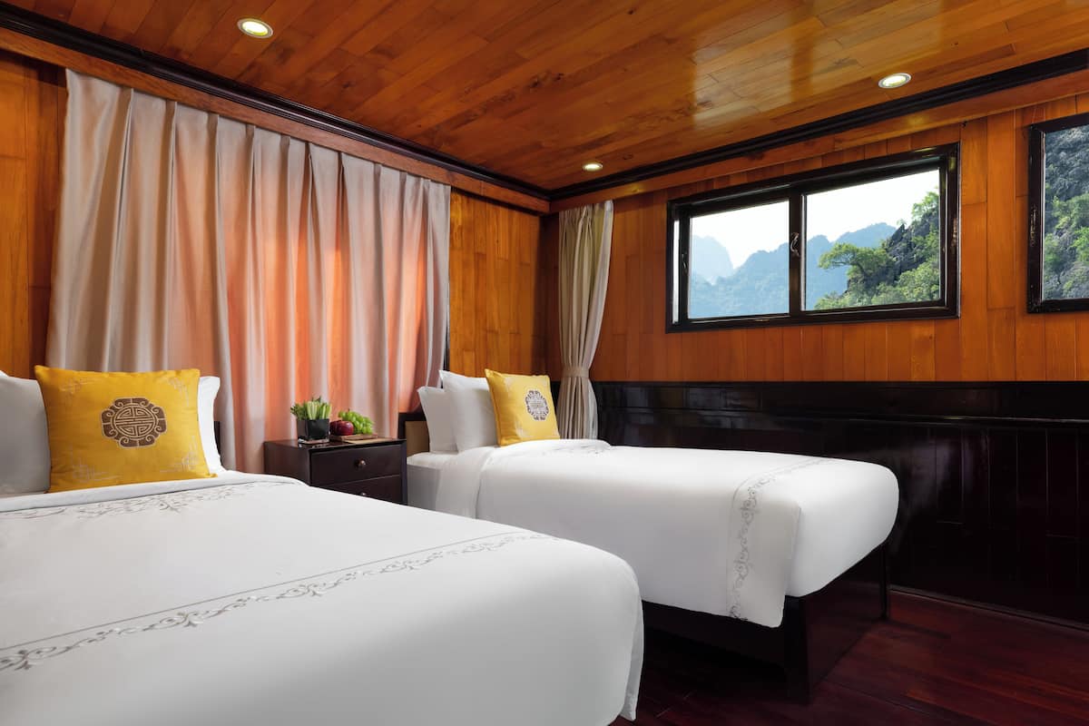 A hotel room with two beds, wooden paneling, a window with a mountain view, and a nightstand with a plant and fruit.
