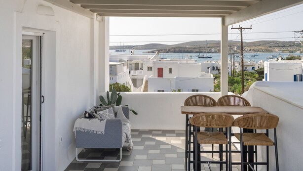 A balcony with a table and chairs, a potted plant, and a view of boats and buildings across the water. - Vagia Calm House 2 (Paros)