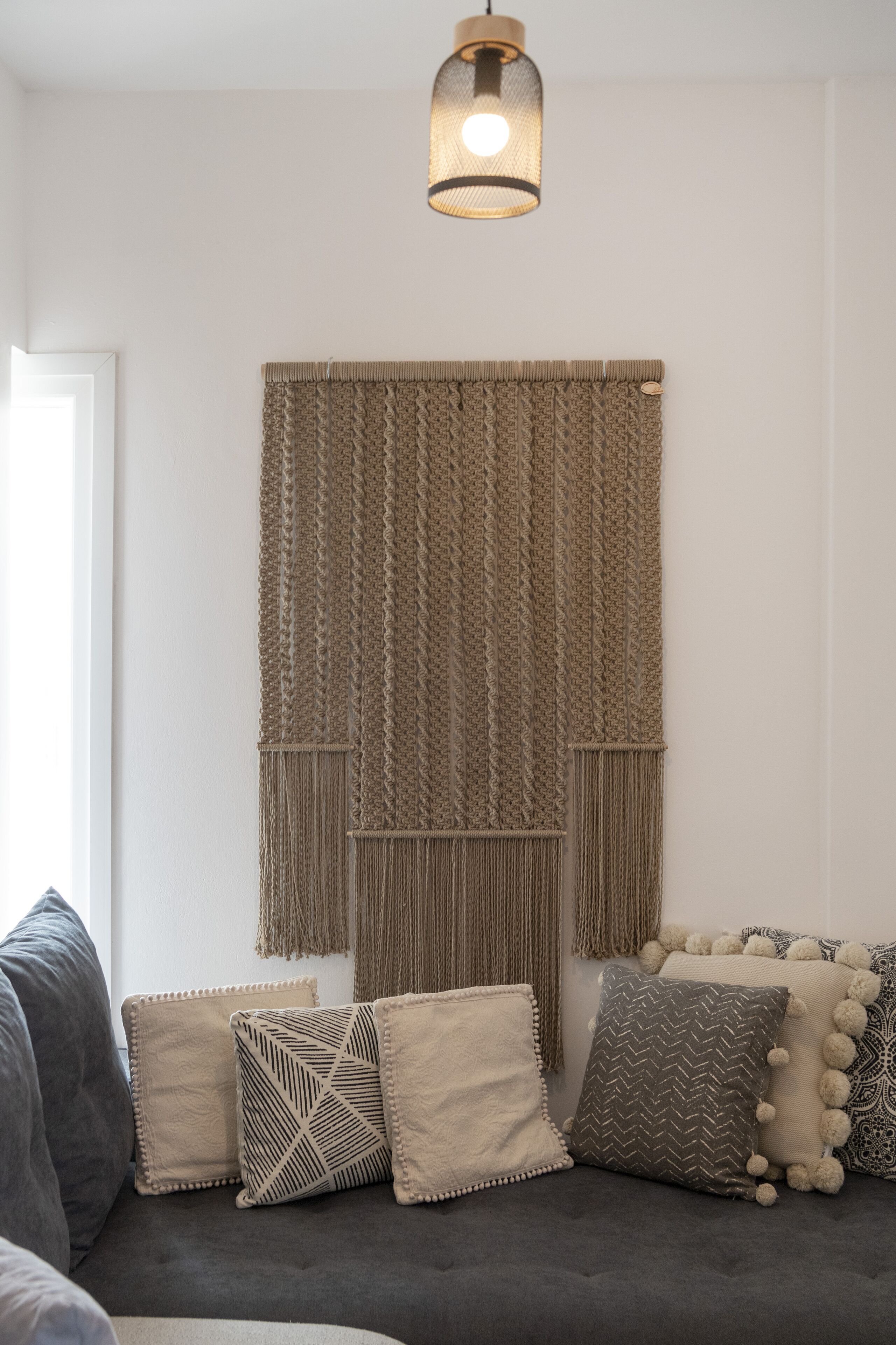 A living room with a grey sofa, a macrame wall hanging, and patterned cushions.