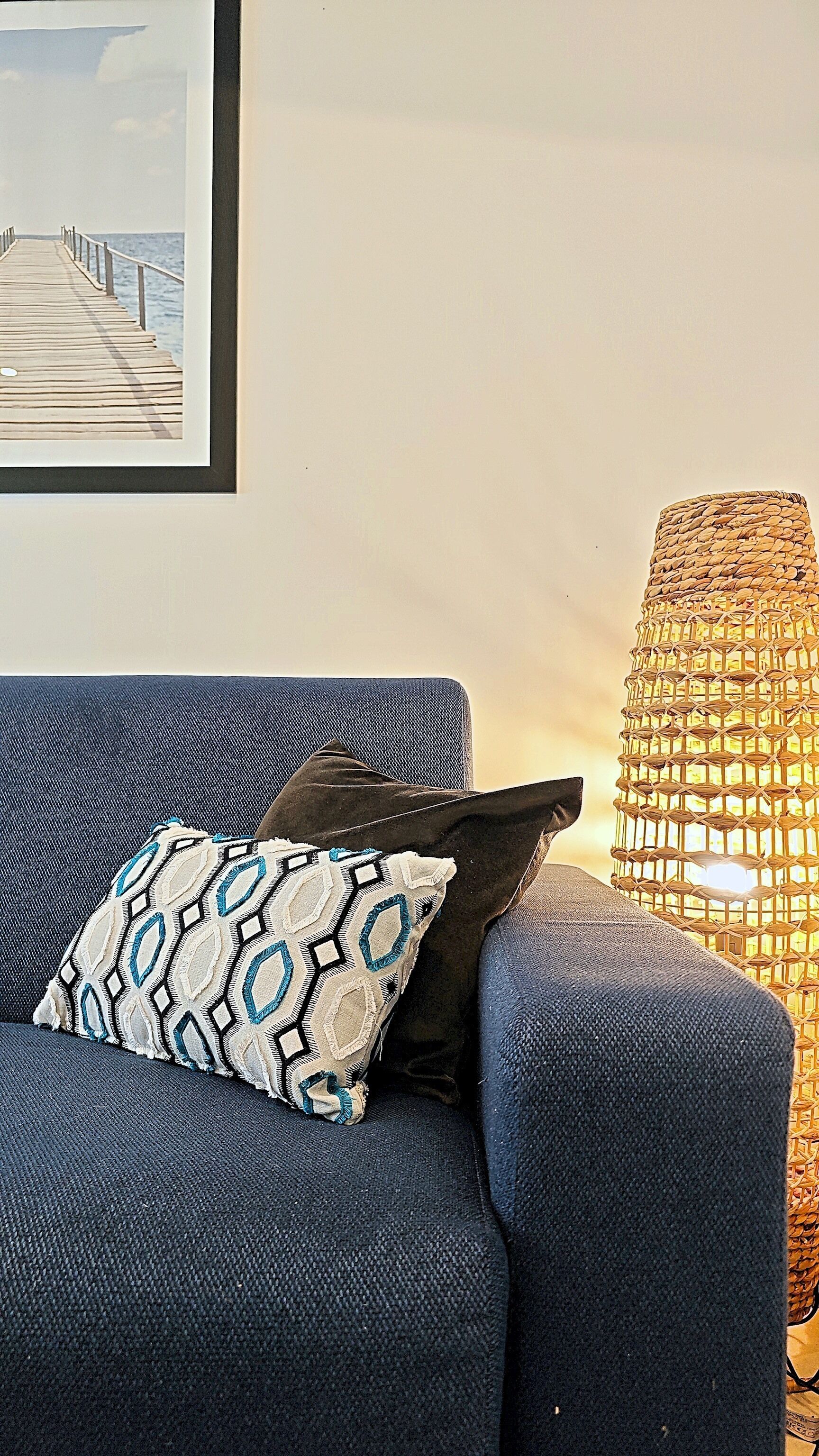 Un divano blu con cuscini decorati, una lampada in vimini e una cornice con l'immagine di un molo.