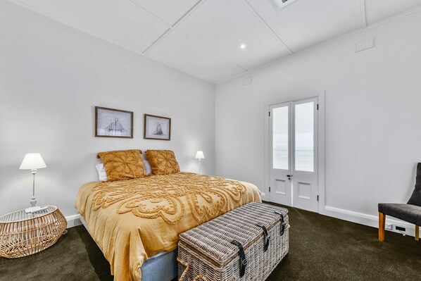A bedroom with a bed, two pillows, a bench, a lamp, a wicker chair, and two framed pictures on the wall.