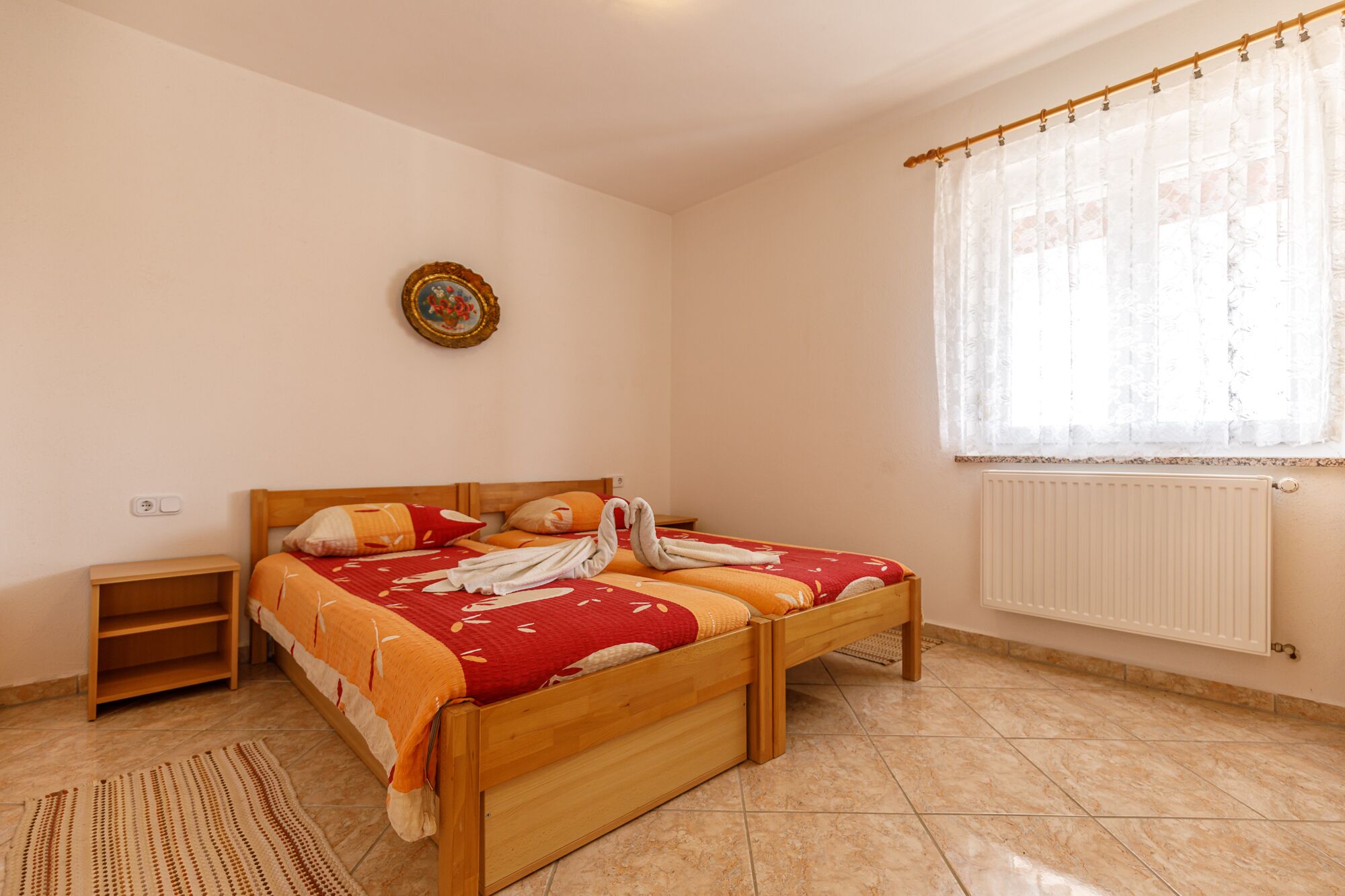 A bedroom with a wooden bed, a bedside table, a radiator, and a window with sheer curtains.