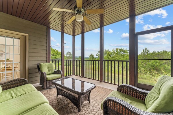 A covered outdoor seating area with wicker furniture and a glass table, overlooking a natural landscape.