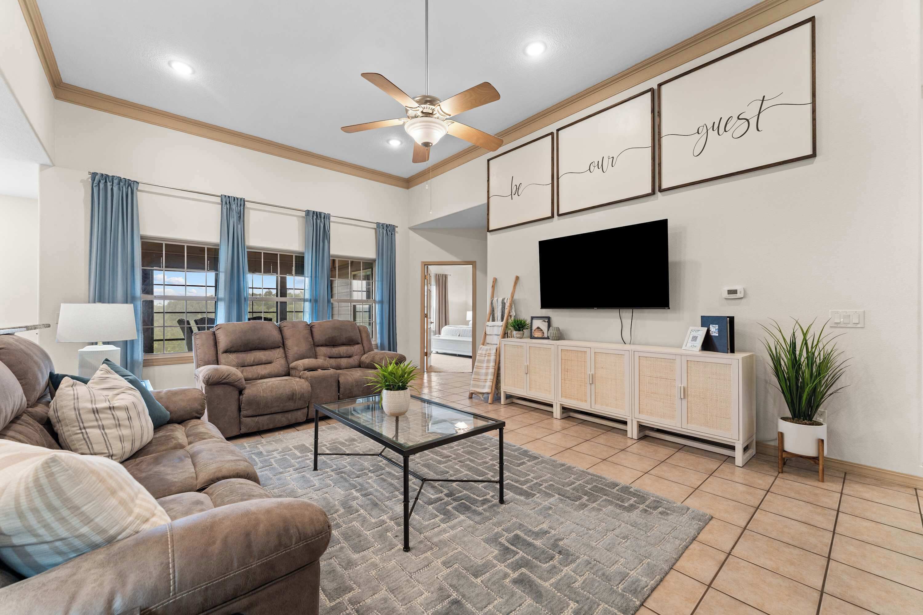 A spacious living room with a ceiling fan, a large flat-screen TV, a sofa set, and a dining area with a buffet cabinet.