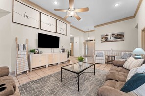 A spacious living room with a flat-screen TV, a ceiling fan, and a large window.