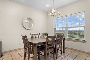 A dining room with a rectangular table, six chairs, a chandelier, a round mirror, and a window with blinds.