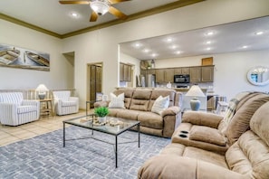 A spacious living room with a large beige sofa, a glass coffee table, and striped armchairs. A kitchen with modern appliances is visible in the background.
