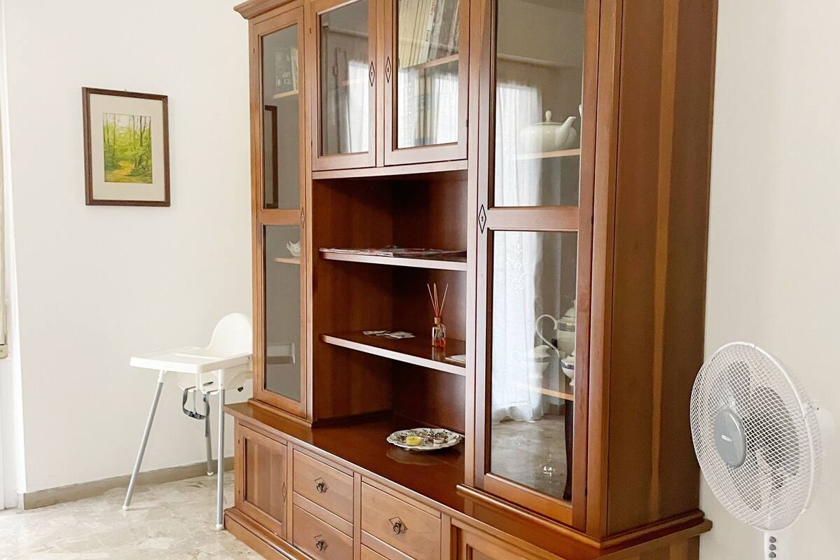 Ein Holzschrank mit Glastüren und Regalen, ein weißer Stuhl und ein Standventilator in einem Raum mit Fliesenboden.