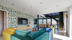 A modern living room with a large sectional sofa, a flat-screen TV mounted on a patterned wall, and a staircase leading to an upper level. - Un nid dans la Grange - Maison avec jacuzzi et sauna (Boué)