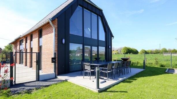 A modern building with a large glass facade, a brick exterior, and a patio area with outdoor seating. - Un nid dans la Grange - Maison avec jacuzzi et sauna (Boué)