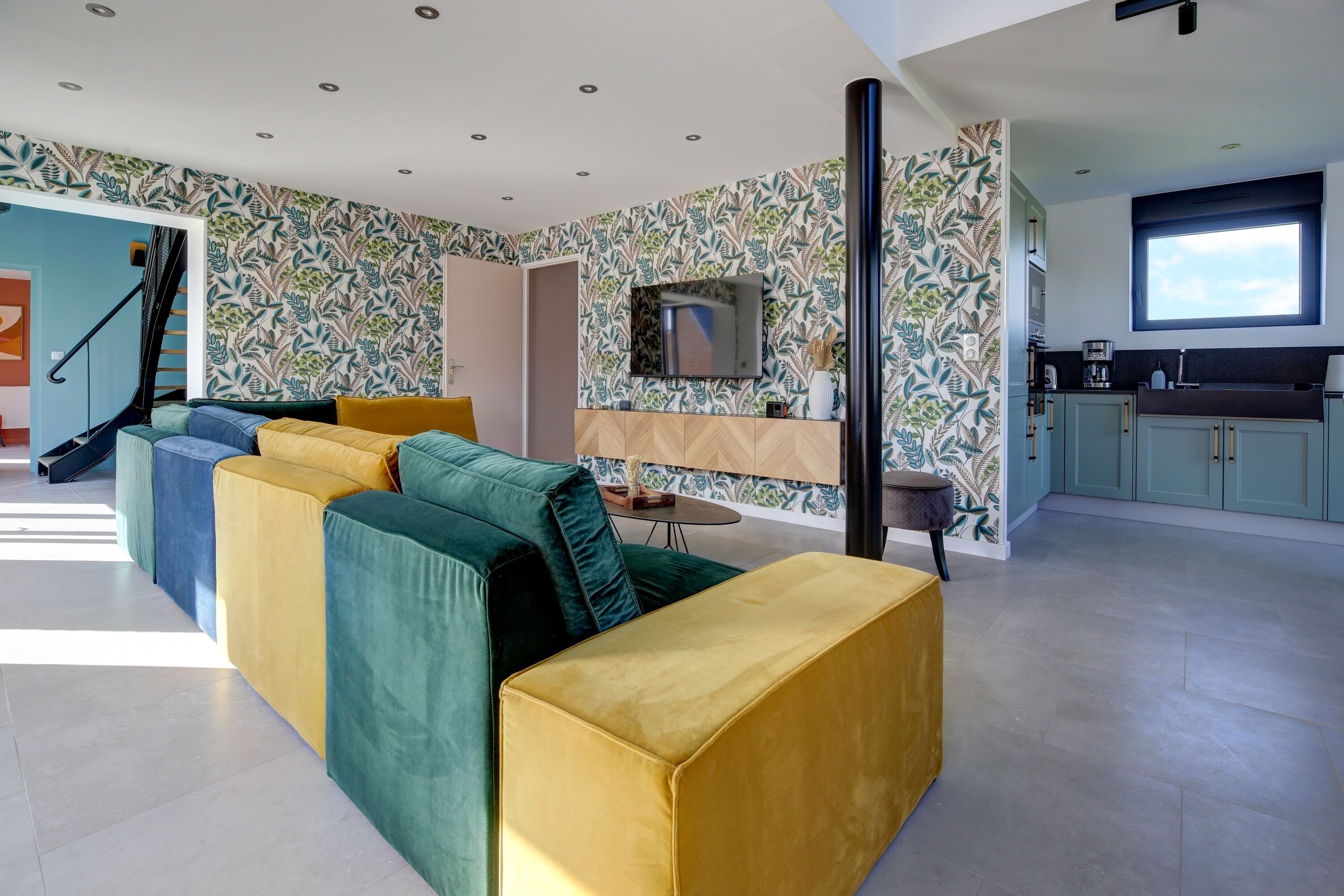 Una sala de estar moderna con un conjunto de sofás, una televisión y una zona de cocina con armarios y un ventanal.