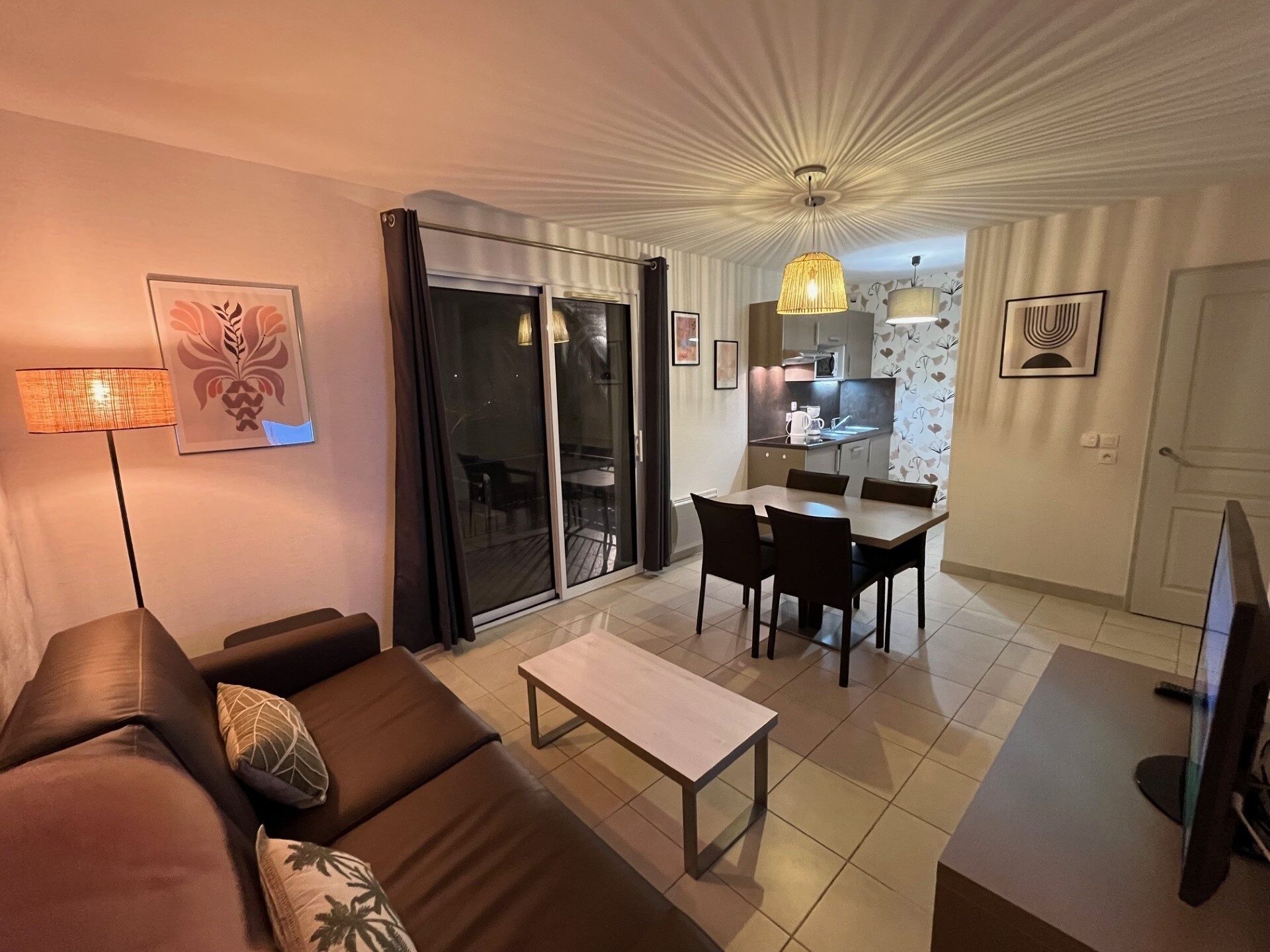 A modern living room with a sofa, a coffee table, and a dining area with a table and chairs. There is a kitchen visible through an open door, and a television is mounted on the wall.