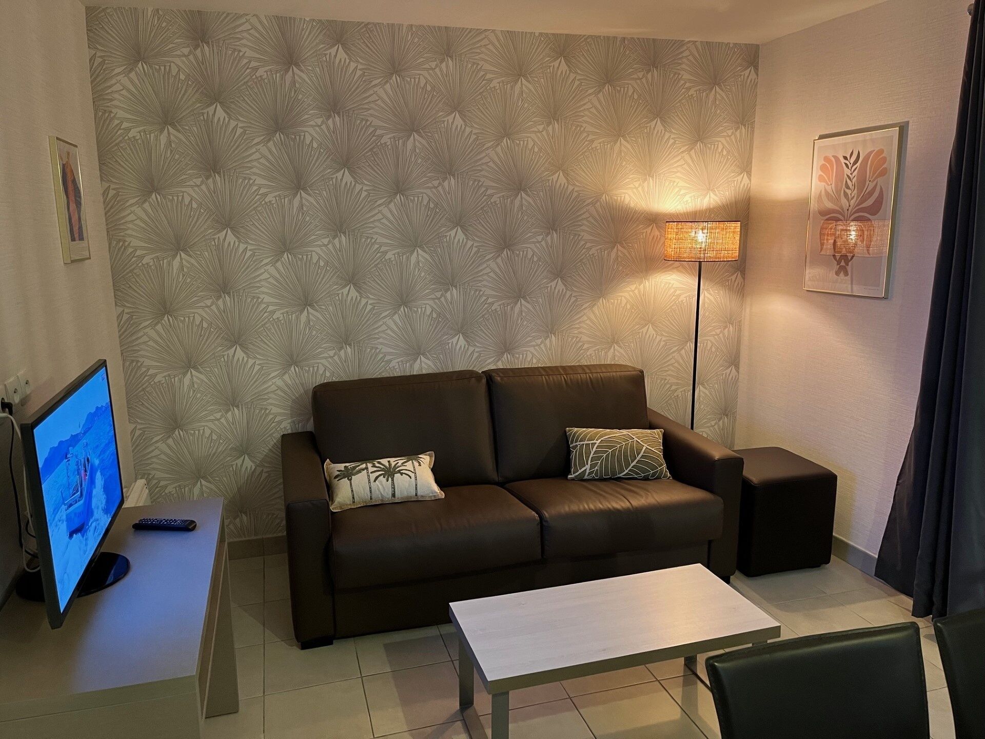 Ein Wohnzimmer mit einem braunen Sofa, zwei Kissen, einem Beistelltisch mit Fernbedienung, einem Fernseher, einer Stehlampe und einem Couchtisch.