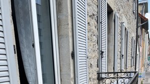 Un balcon avec une table et des chaises, un bâtiment en pierre avec des volets, et un ciel dégagé.