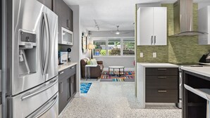 Een moderne keuken met roestvrijstalen apparatuur, een eetgedeelte met een kleurrijk tapijt, en een woonruimte met een bank en ramen.