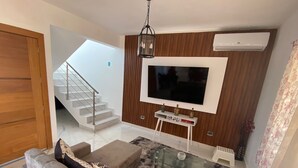 Een moderne woonkamer met een bank, een glazen salontafel, een aan de muur bevestigde televisie, en een trap die naar een hoger niveau leidt.