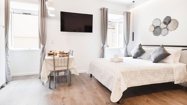A bedroom with a bed, a TV, a dining table with chairs, and a window with curtains. - Saint's View Apartments (Corfu)