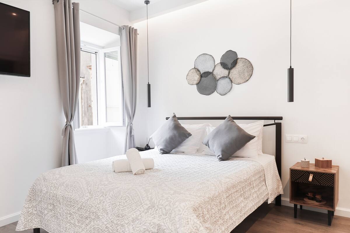 Ein ordentlich bezogenes Bett mit einem gemusterten Bettbezug, ein Wandbild und ein Nachttischchen mit einer kleinen Lampe.