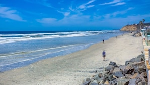 Ein Strand mit felsigem Ufer, blauem Himmel und ein paar Personen, die am Strand entlanggehen.