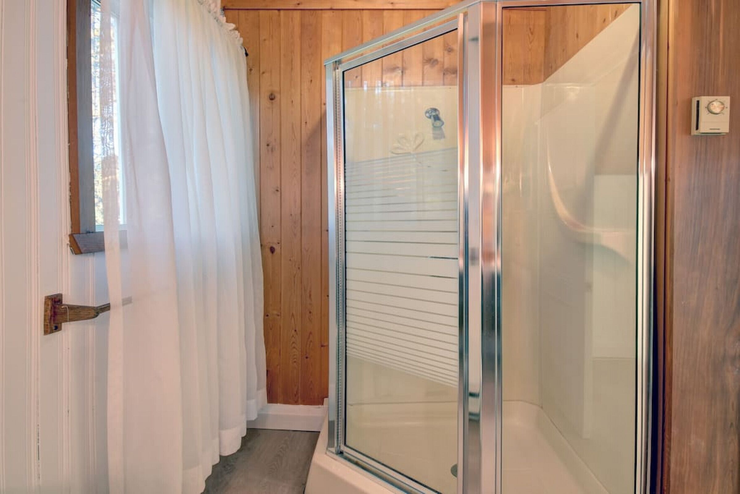 A bathroom with a glass shower enclosure, wooden walls, and a white door with a curtain.