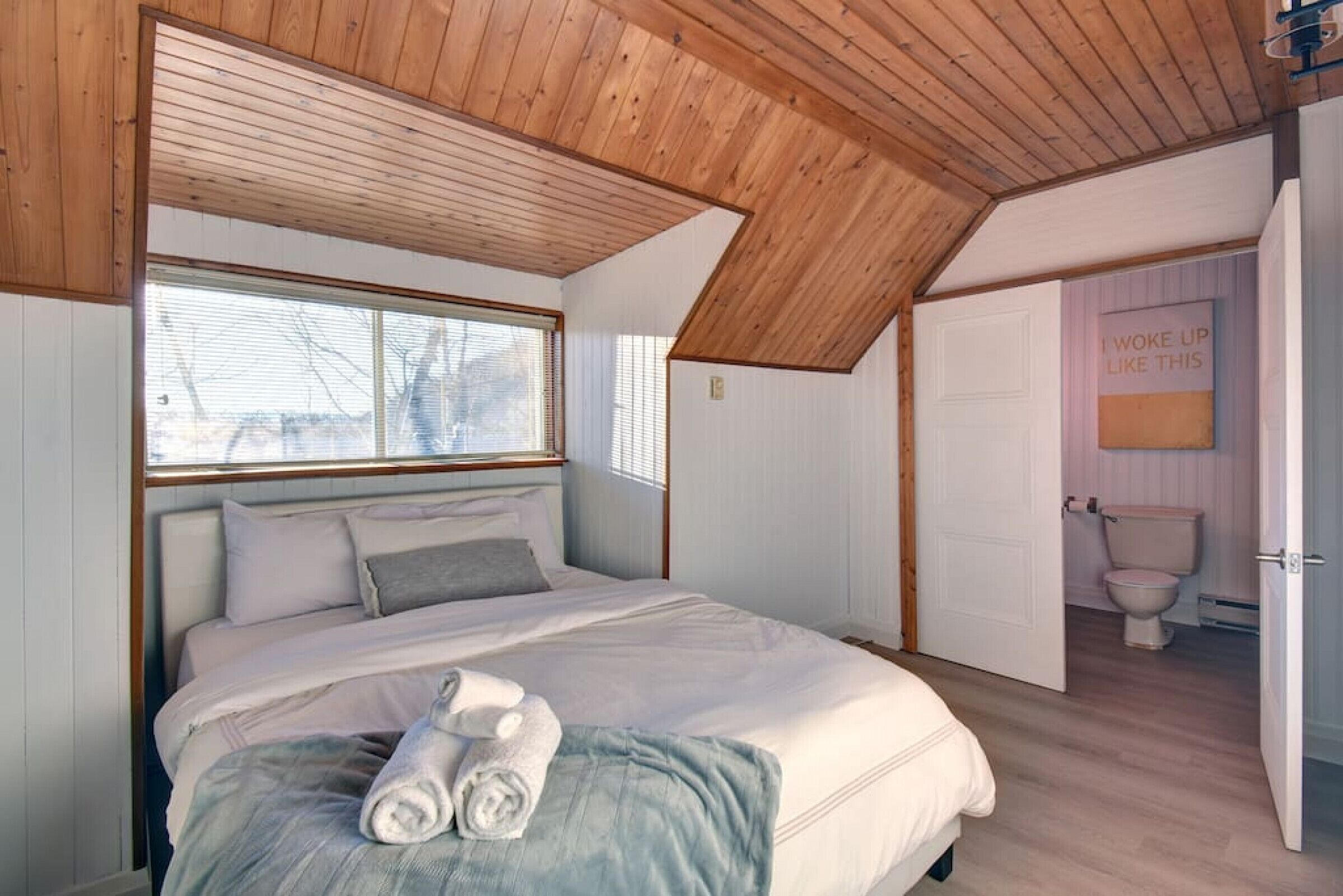 A bedroom with a sloped ceiling, a large window, a bed with white linens, and a toilet in the adjacent room.