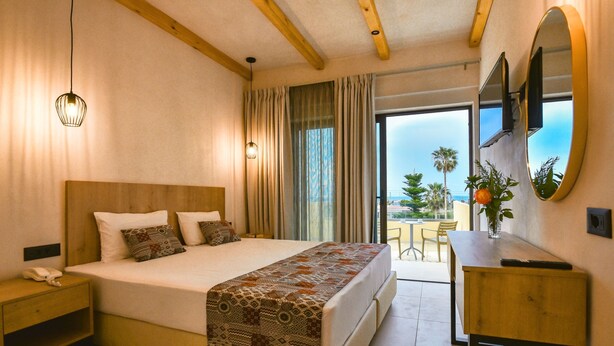 Habitación de hotel con una cama grande, mesita de noche y un escritorio con silla. Se ve una ventana con cortinas y vistas a palmeras.