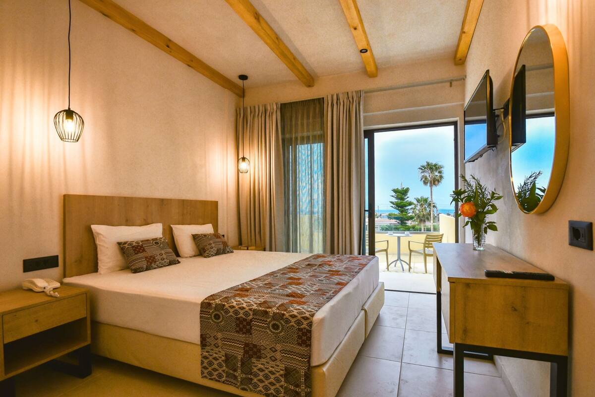 Habitación de hotel con una cama grande, mesita de noche y un escritorio con silla. Se ve una ventana con cortinas y vistas a palmeras.