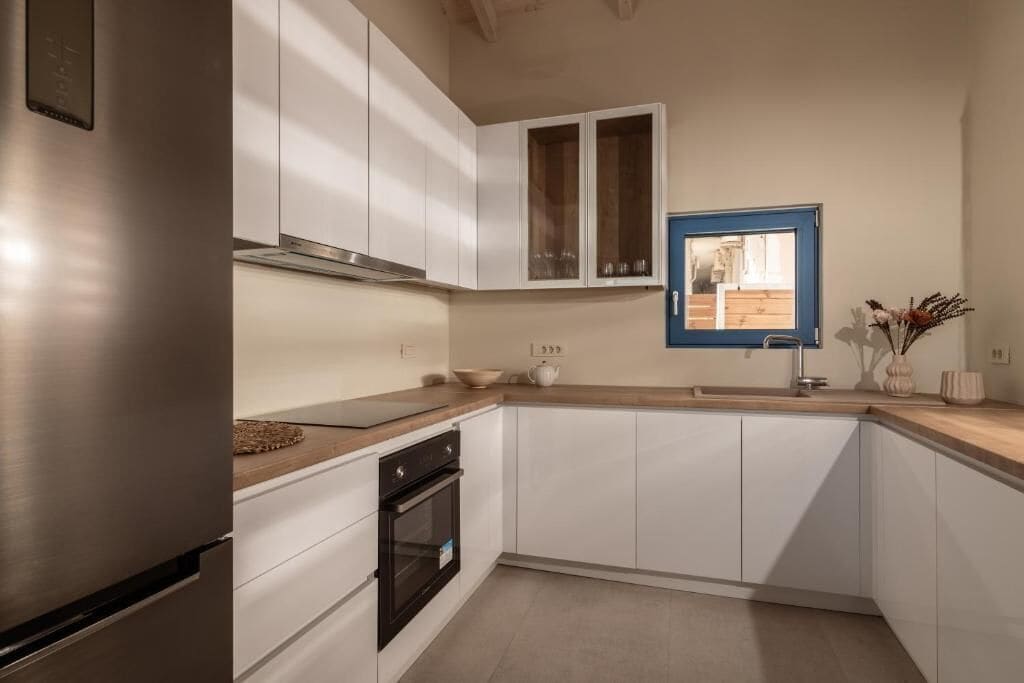 A modern kitchen with white cabinets, a stainless steel refrigerator, a built-in oven, and a window with a view of the outdoors.