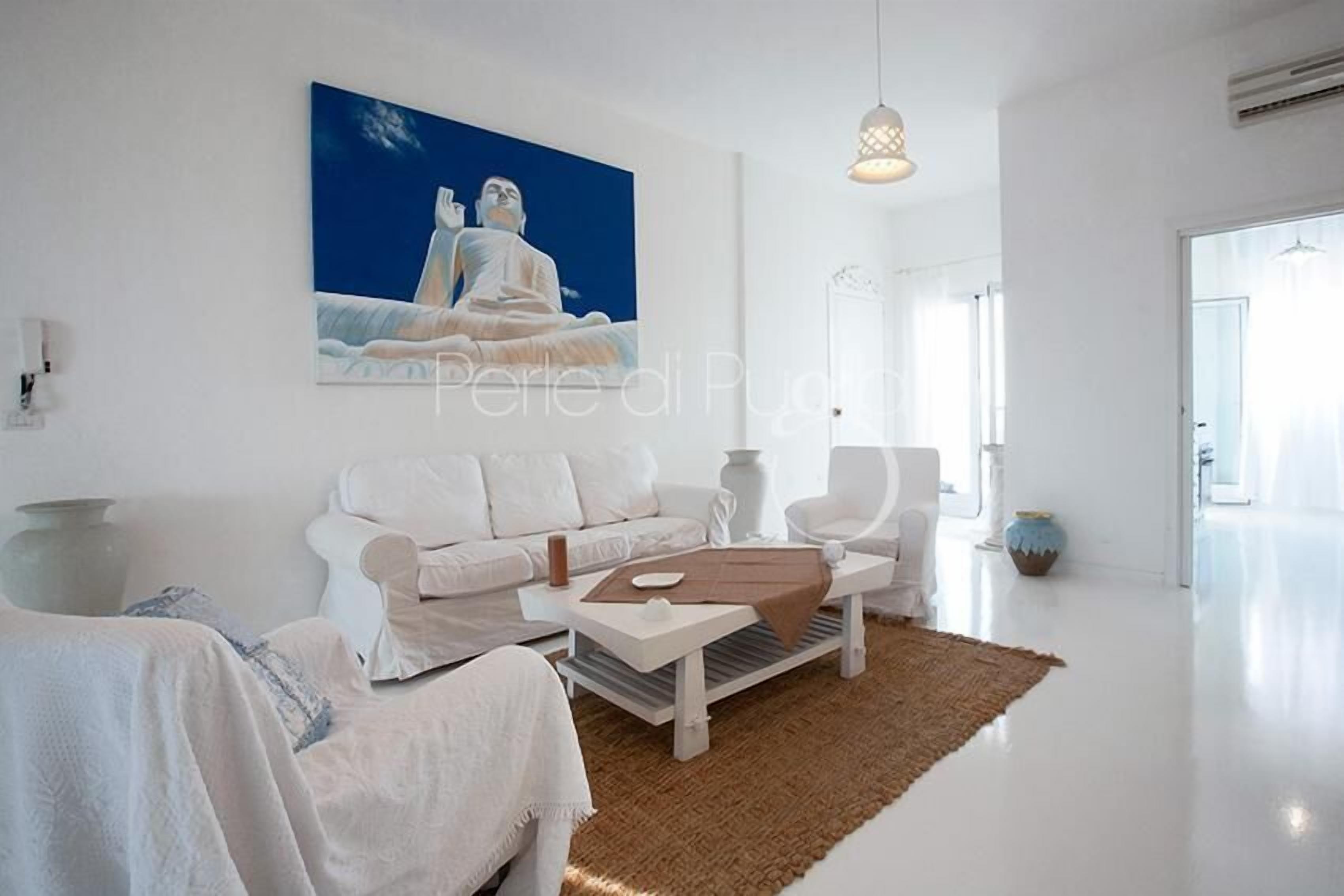 A spacious living room with a white sofa, a coffee table, and a painting of a meditating figure.