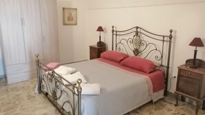 Uma cama bem arrumada com estrutura de metal, uma mesa de cabeceira de madeira com abajur, e um guarda-roupa num quarto com parede lisa e um quadro emoldurado.