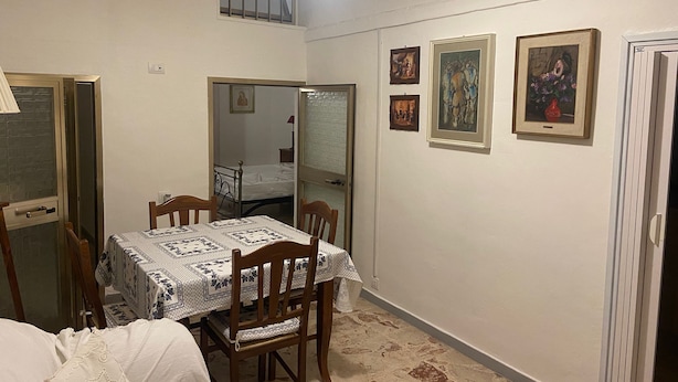 Quarto pequeno e bem iluminado com uma mesa de jantar, cadeiras e uma cama. Há quadros nas paredes e uma janela com grades.