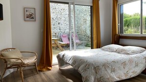 A bedroom with a bed, a chair, a window, and a view of an outdoor area with a stone wall and chairs. - Maison de campagne, Sainte Mère Église, site historique (Sainte-Mère-Eglise)