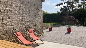 A stone wall, two red lounge chairs, a wooden table, and two potted plants on a gravel surface. - Maison de campagne, Sainte Mère Église, site historique (Sainte-Mère-Eglise)