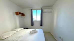 Una habitación pequeña y limpia con una cama individual, una ventana con cortinas, un equipo de aire acondicionado y un estante de madera.