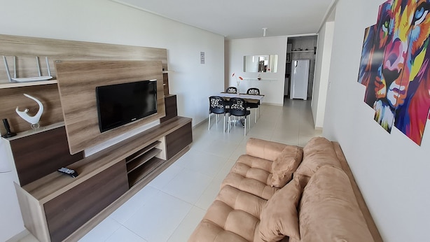 Una sala de estar moderna con televisor de pantalla plana, mueble de madera para entretenimiento, sofá color beige, comedor con sillas y una cocina al fondo.