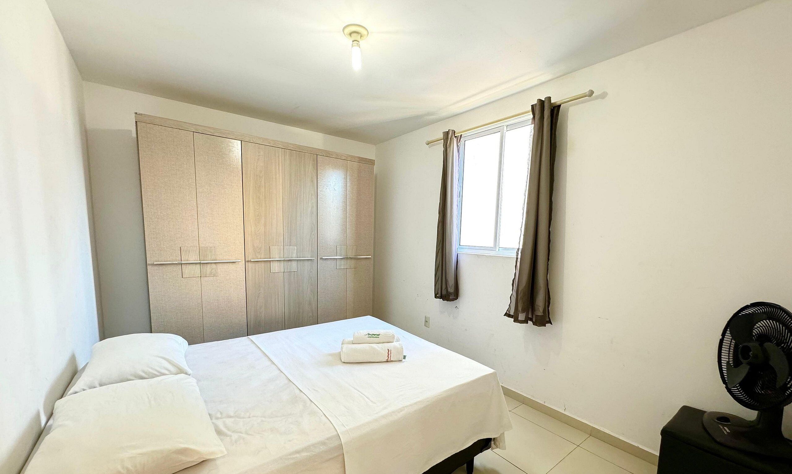 Un dormitorio con cama, armario, ventana con cortinas y un ventilador de pie.